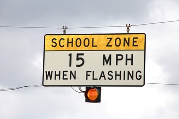 School Zone 15 Miles Per Hour When Flashing Yellow and White Sign with Black Lettering Hanging on Cables with Orange Light Below Illuminated Sunlit against Cloudy Sky in the Afternoon