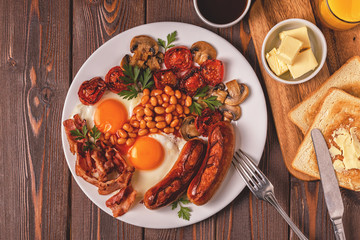 Traditional full English breakfast with fried eggs, sausages, beans, mushrooms, grilled tomatoes and bacon on wooden background.