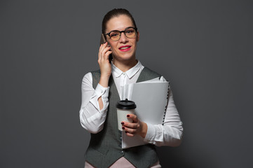 Beautiful young businesswoman in formal suit talking on smartphone and coffee mug standing against gray background