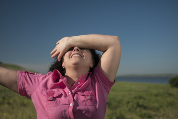brunette in a pink shirt smiles and covers her face with her hands in nature against a blue sky