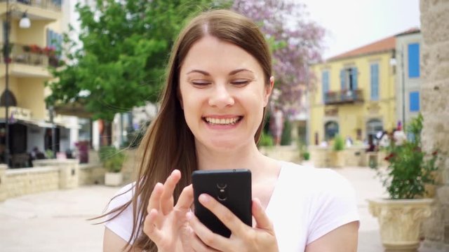 Slow Motion. Smiling young woman in white t-shirt walking down street in european city using cellphone. Happy female browsing, reading news, chatting with friends via mobile phone