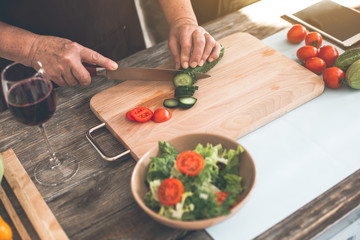 Top view close up of elderly female hands making salad in kitchen. Woman is cutting a cucumber by knife on board