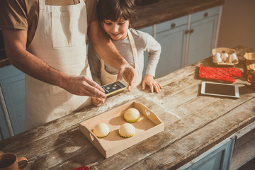 Father and child standing in kitchen and making snapshot of sweets on tray. Copy space in right side