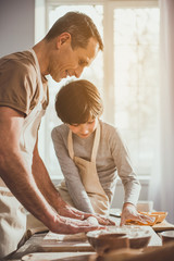 Father standing in apron and unrolling soft dough on table while his kid observing process. They are smiling