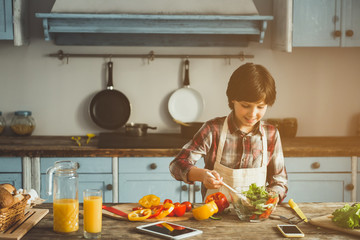 Happy child in apron standing in kitchen and mixing vegetable salad. Gadgets and juice are on desk