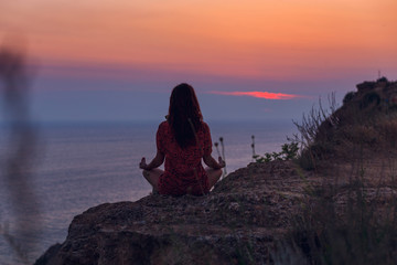 Meditation on the beach. A woman sits on a cliff above the ocean and does yoga. Femininity. Force. Soft shades at sunset. Beautiful scenery. Amazing sunset on the sea. Cloudy sky.Magic of nature.