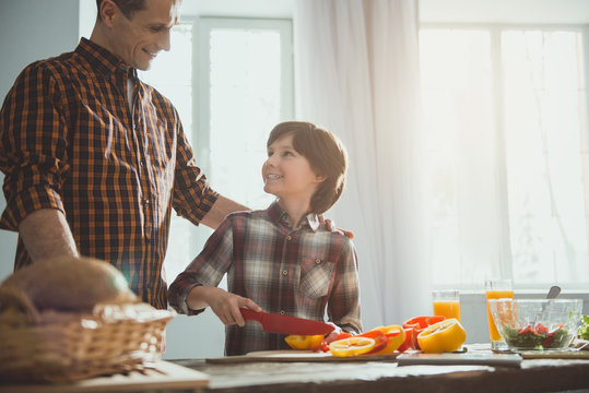 Man And Boy Standing In Cooking Room. Child Slicing Veggies While Dad Looking At Him With Proud. Copy Space In Right Side