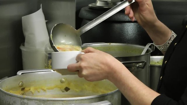 Woman in restaurant kitchen ladles soup into bowl and serves with bread roll and butter