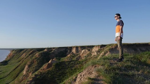 Profile of a snappy man in a baseball cap, sunglasses and a T-shirt standing on the Black Sea shore and looking at seascape in summer in slow motion