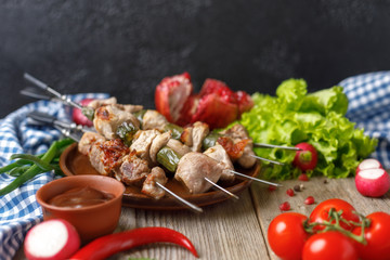 Juicy delicate kebabs of pork on skewers laid out on a dish and fresh vegetables. Still life on a wooden background with textiles. Close-up.