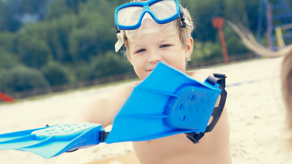 Little Caucasian boy with snorkeling equipment sitting on sand on tropical beach and playing with flippers on hands.