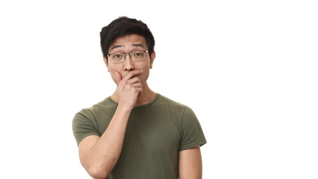 Portrait of concerned asian man 20s wearing glasses being shocked with unexpected news and covering open mouth in anxiety, isolated over white background. Concept of emotions
