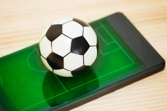 A toy soccer ball on a smartphone with a picture of a green field. Concept of the game of football.