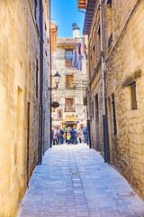 Fototapeta na wymiar Laguardia, Alava, Spain. March 30, 2018: Narrow cobblestone street with facades of masonry stone houses and union with the main street of the town, full of people