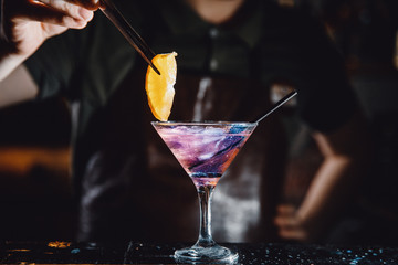 Barman prepares cocktail with orange and martini purple color on bar with alcohol. Uses tongs for decoration. Dark background.