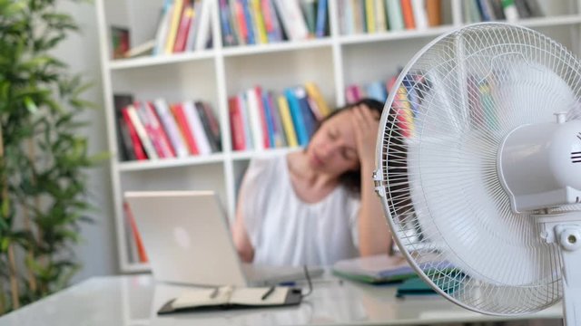 The video is about one businesswoman feeling hot and trying to refresh in summertime heat at work in office.Shot fixed on the electric fan and the woman acts defocused on the background.