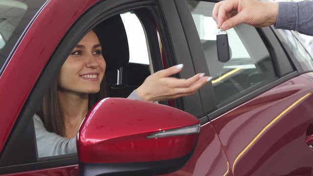 Cheerful young attractive woman sitting in her newly bought automobile smiling happily receiving car keys from the salesman at the delaership showroom. Shopping, travel, ownership, driving.