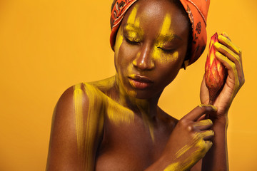 Cheerful young african woman with yellow makeup on her eyes. Female model against yellow background with orange flower.