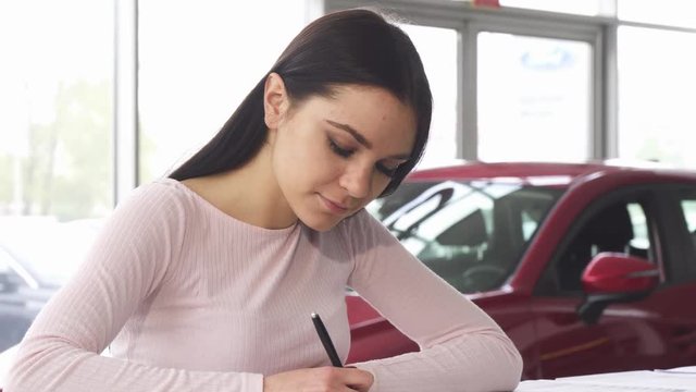 Gorgeous young woman smiling signing papers while buying a new car at the dealership salon. Attractive female customer filling documents after purchasing an auto. Insurance, sales concept.