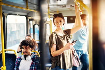 Young beautiful woman is standing in the bus using phone and holding onto the bar while waiting to...