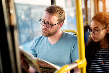 A young adorable couple is sitting together on a bus seat and reading newspapers as they wait for...