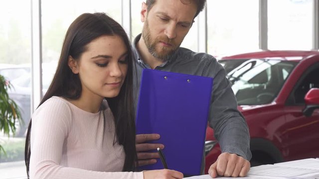 Professional car salesman helping his female client signing papers after buying a new car at the dealership showroom. Beautiful woman filling documents for her newly bought auto. 