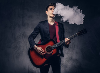 Obraz premium Handsome young musician with stylish hair in elegant clothes exhales smoke while playing acoustic guitar.