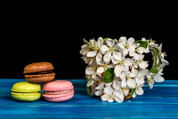 Multicolored macaroons and cup decorated with white flowers of apple trees on a blue wooden background.