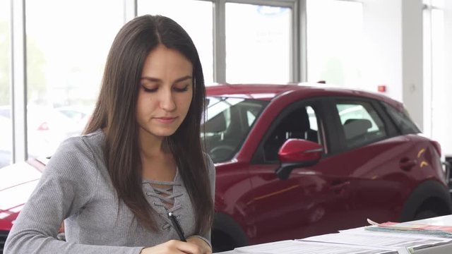 Attractive woman buying a new car at the dealership and signing papers for insurance. Young female customer filling documents for her new automobile. Consumerism, ownership concept.