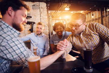 Exhausted and focused male friends having arm wrestling challenge at the local bar while other supporting.