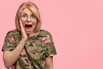 Horizontal shot of stunned blonde female exclaims with horrified expression as watches horror film,...