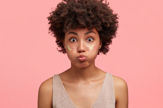 Funny amazed attractive African American female blows cheeks, has bated breath, cheeks decorated with spangles, stares at camera, prepared for party with friends, isolated over pink background