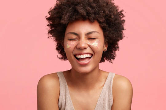 People, happiness, positive emotions concept. Satisfied dark skinned woman with curly bushy hair, laughs joyfully, keeps eyes shut, has white perfect teeth, hears funny story or comic anecdote