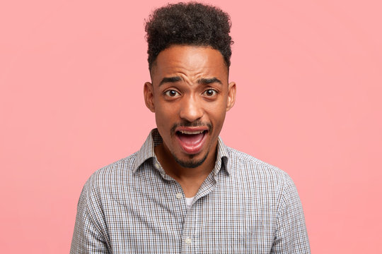 Attractive dark skinned amazed male student has discontent expression, doesn`t agree with mark, argues with professor, isolated on pink background. Emotional African American hipster guy indoor