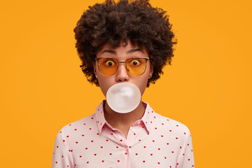Playful funny African American woman blows chewing gum, spends spare time with friends, wears shirt...