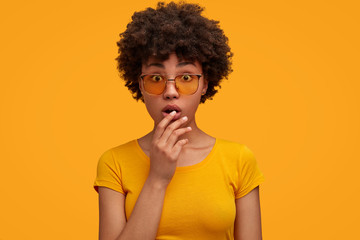 Horizontal shot of surprised African American female in bewilderment, feels shocked, keeps mouth opened, listens to exciting story, isolated over yellow background. Bright warm colours prevail