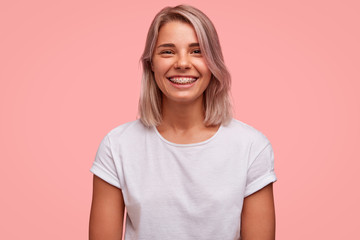 Portrait of happy female with broad smile, wears brackets on teeth and casual white t shirt, poses against pink background. Attractive woman with bobbed hairstyle, being in good mood after date
