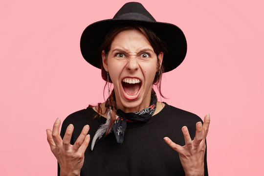 Portrait of irritated young cowgirl gestures angrily, shows annoynace, being mad and crazy, yells with negativity, opens mouth widely, wears black stylish clothes, isolated over pink background