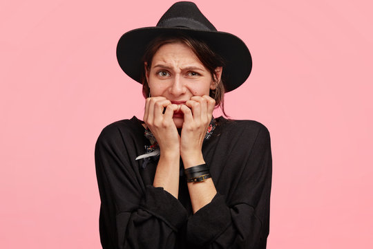 Photo of nervous displeased European woman bites finger nails, worries before presentation of her project, wears black hat, poses against pink studio background. Negative human facial expressions