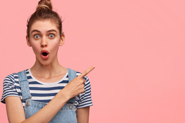 Pretty lovely European female with stunned expression, indicates aside at balnk copy space for your advertisement, wears casual t shirt, poses against pink background. People, style conncept