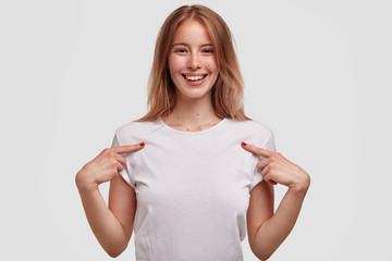 Happy glad attractive woman with lovely look, broad shining gentle smile, indicates at t shirt on...