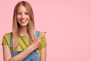 Horizontal portrait of positive European woman shop assistant advertises new product and announces about big discount, points at blank pink background for your promotional text or advertisement