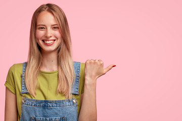 Photo of delighted smiling female student being in good mood, rejoices successfully passed exam, points with thumb at blank copy space for your advertisement. People, positive emotions concept