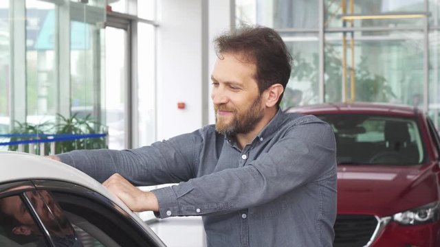 Cheerful mature handsome bearded man examining a new automobile at the dealership showroom. Happy attractive male customer smiling joyfully posing with a new car.
