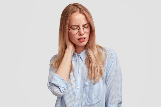 Tired female office worker in spectacles has pain in neck as sat long time in front of computer and prepared report, looks fatigue and exhausted, suffers from ache, isolated on white background.