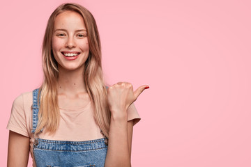 Horizontal shot of delighted attractive female seller advertises her product, indicates at blank copy space for your hearder or promotional text. Successful woman distance worker gestures indoor