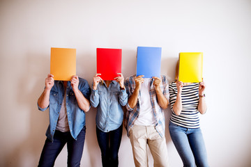 Group of young stylish people holding colourful folder in front of their heads while leaning against the wall in the waiting room for the job interview.