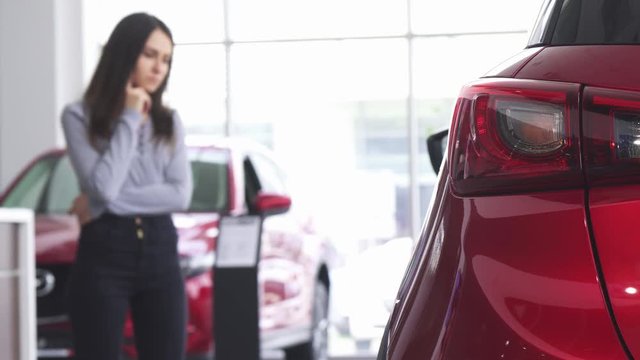 Selective focus on car lights on the foreground. Female customer examining cars for sale on the background. Woman shopping for a new vehicle. Buying cars concept. Driving automobile.