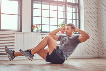 Handsome fitness man in a t-shirt and shorts doing abdominal exercises on floor at home.