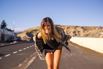 Fototapeta na wymiar cute model beautiful blonde long hair smile to you in camera walking on a long road to travel with a skateboard behind. mountains background and sunlight day of vacation in tenerife